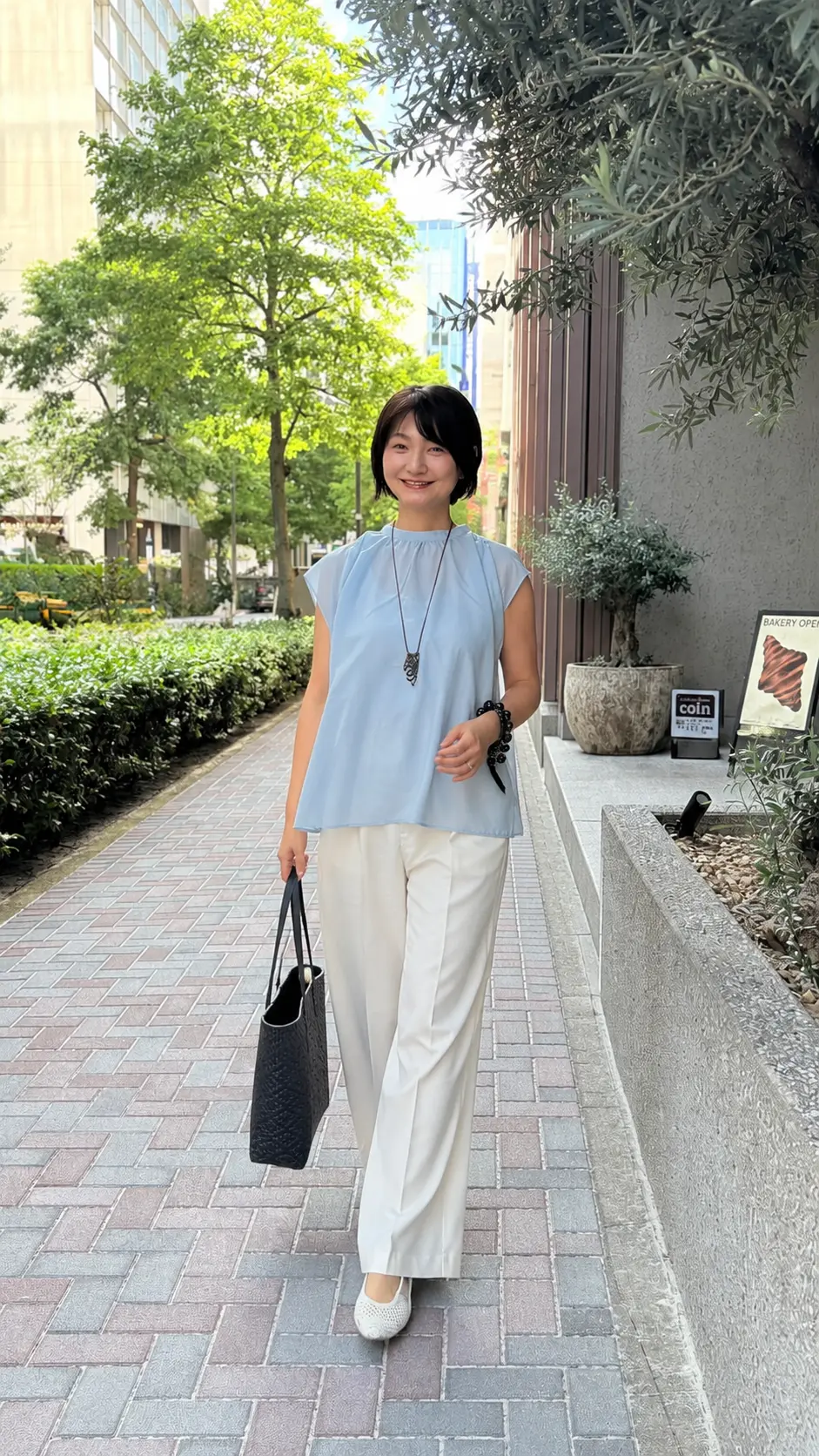 青色のネックラインが特徴的なトップスに白いパンツを合わせた女性が、石畳の道で微笑みながら立っている。黒いバッグを持ち、腕にはブレスレットを身に着けている。背景には緑豊かな植栽と建物が見える。