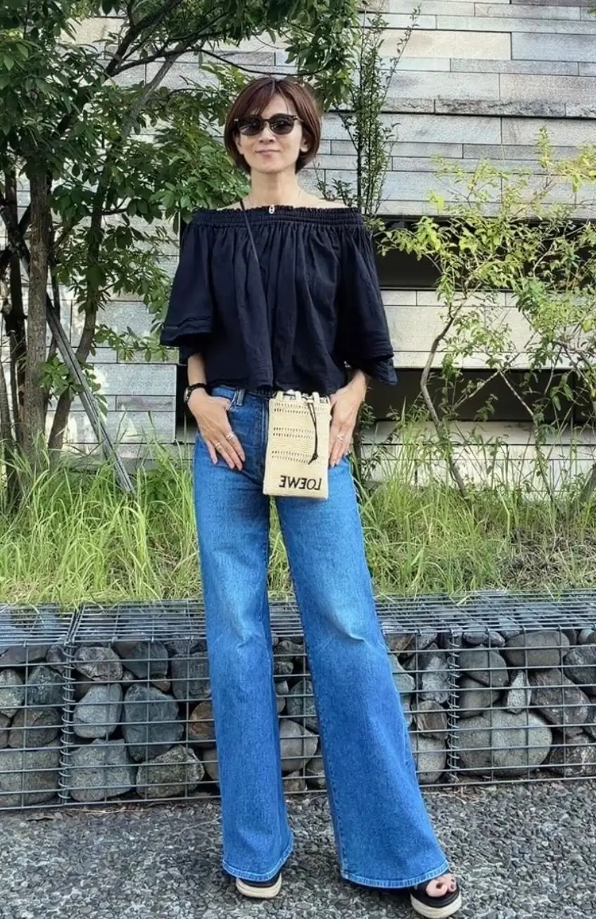 黒いオフショルダーのトップスに、濃い青のフレアジーンズを合わせた女性。茶色のサングラスをかけ、TOUMEの黄色のミニバッグを斜め掛けしている。背景には緑の木々と石の壁がある。
