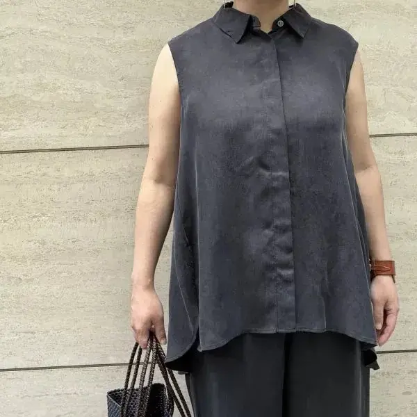 グレーのノースリーブシャツを着た女性。黒いパンツを着用し、茶色のバッグと腕時計を持つ。背景はベージュの壁。