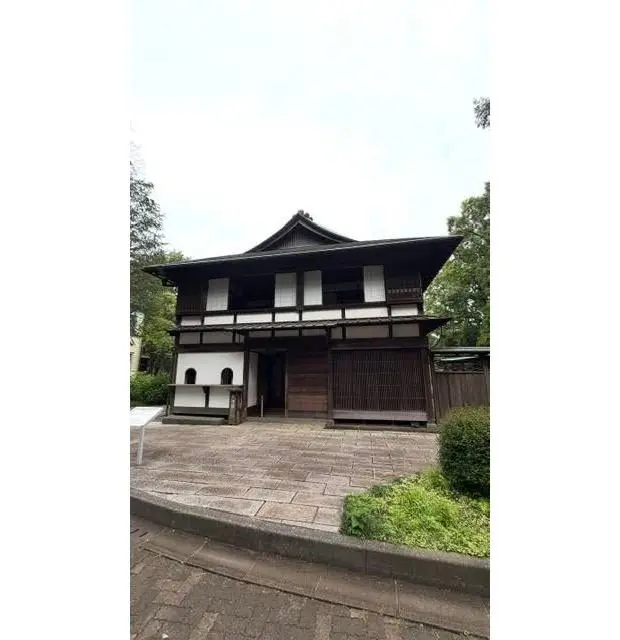 伝統的な和風建築の二階建ての家、木造の外観と白い壁、窓と扉が備えられた入り口、石の敷地と低木が周囲に配置