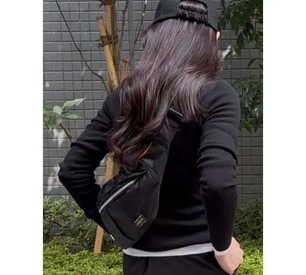 黒いニットにキャップをかぶった人物の後ろ姿。茶色の長い髪が肩にかかり、PORTERの黒いボディバッグを斜め掛けしている。背景にはグレーのタイル壁と緑の植栽がある。