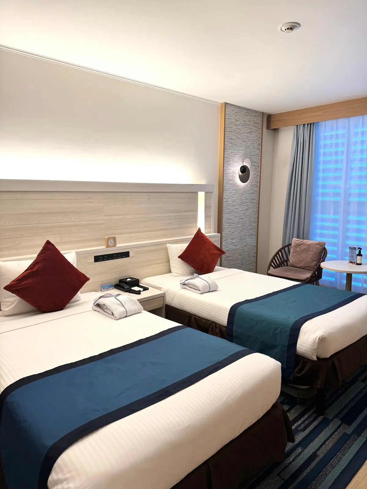 ホテルの客室。2台のシングルベッドが並び、それぞれに白いシーツと青色の装飾が施されている。枕には赤色のクッションが置かれ、壁面には間接照明がある。窓のカーテンは青色で、部屋全体が清潔感のある雰囲気。