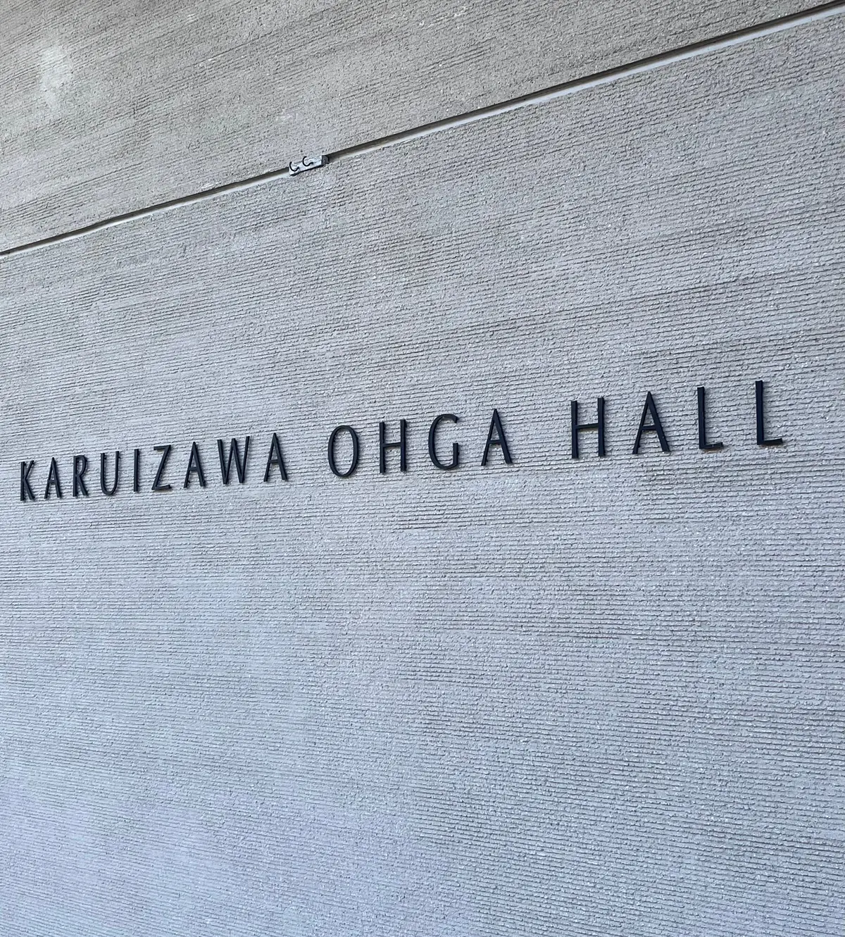 灰色のコンクリート壁に、縦の溝が施されている。中央に黒色の文字で「KARUIZAWA OHGA HALL」と刻まれている。