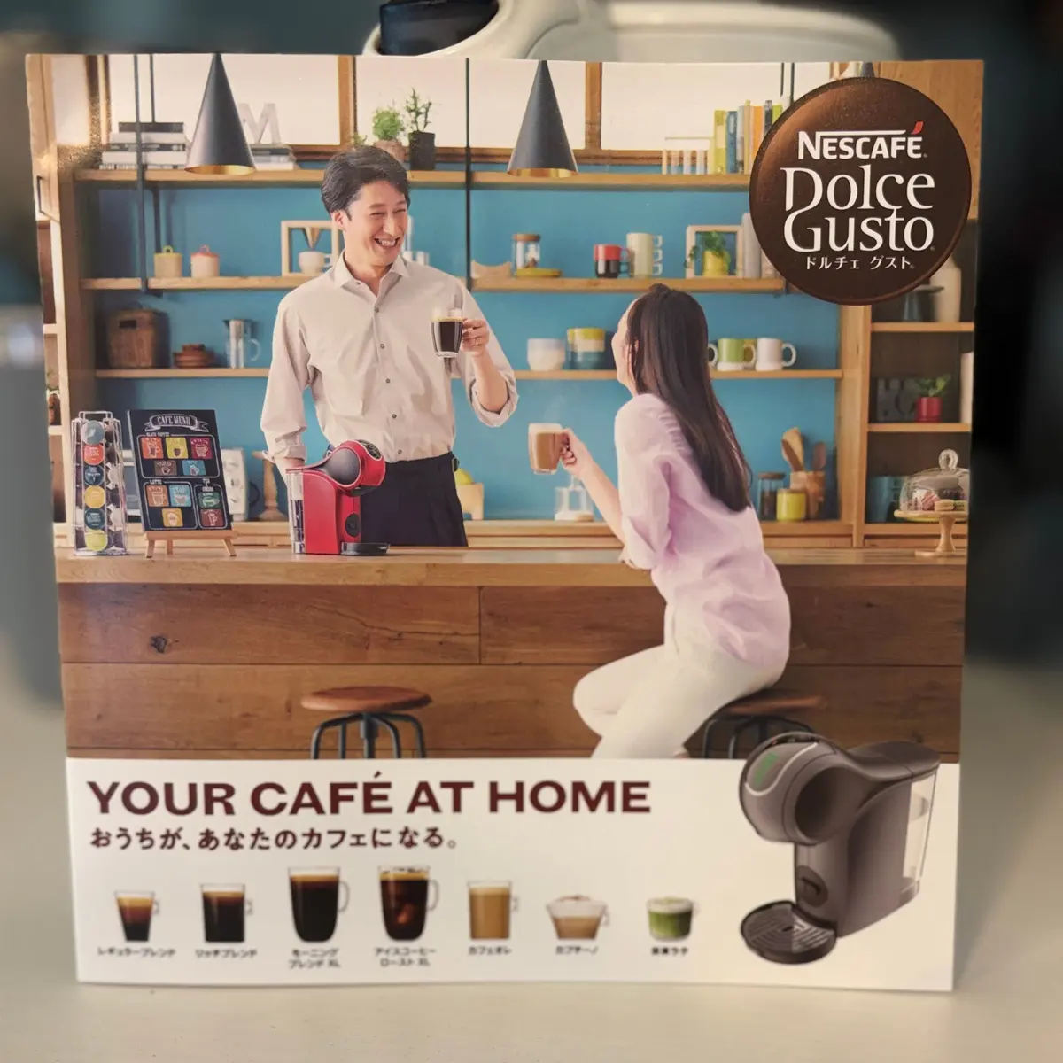 ネスレ ドルチェ グストの広告。カフェのような空間で、男性が女性にコーヒーを注いでいる様子を写した写真。下には「YOUR CAFÉ AT HOME」の文字と、様々なコーヒーカップのイラストが並んでいる。