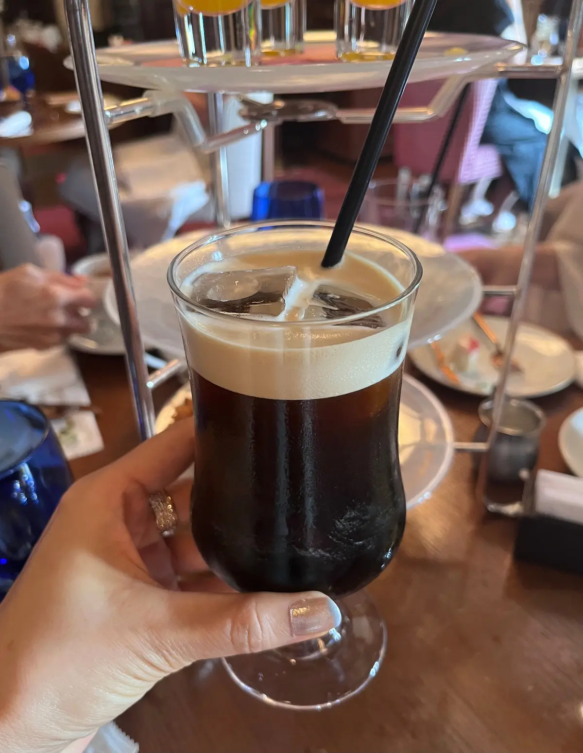 カクテルグラスに入った、黒色の液体と泡が層になっている飲み物。氷が浮かび、ストローが挿さっている。木製のテーブルに置かれ、背景にはレストランの様子が見える。