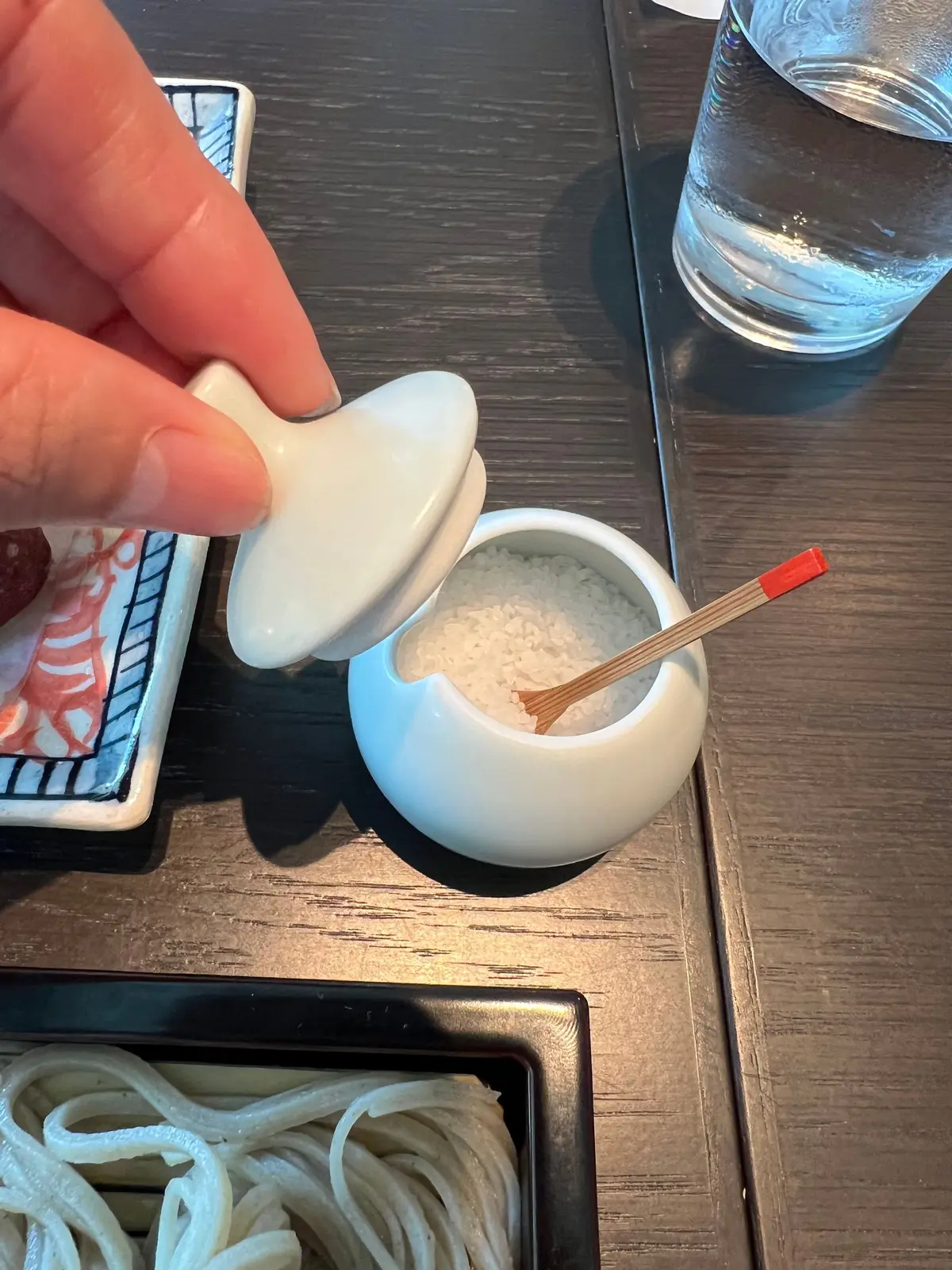 白い陶器の塩入れから塩をすくい、木製の塩さじで取り出す様子。木製のテーブルに水の入ったグラスと、うどんが盛られた器も見える。