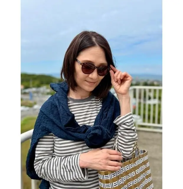 サングラスをかけた女性が、ストライプの長袖シャツに濃い青色のスカーフを羽織り、籐製のバッグを持っている。背景には海と緑の丘が見える。