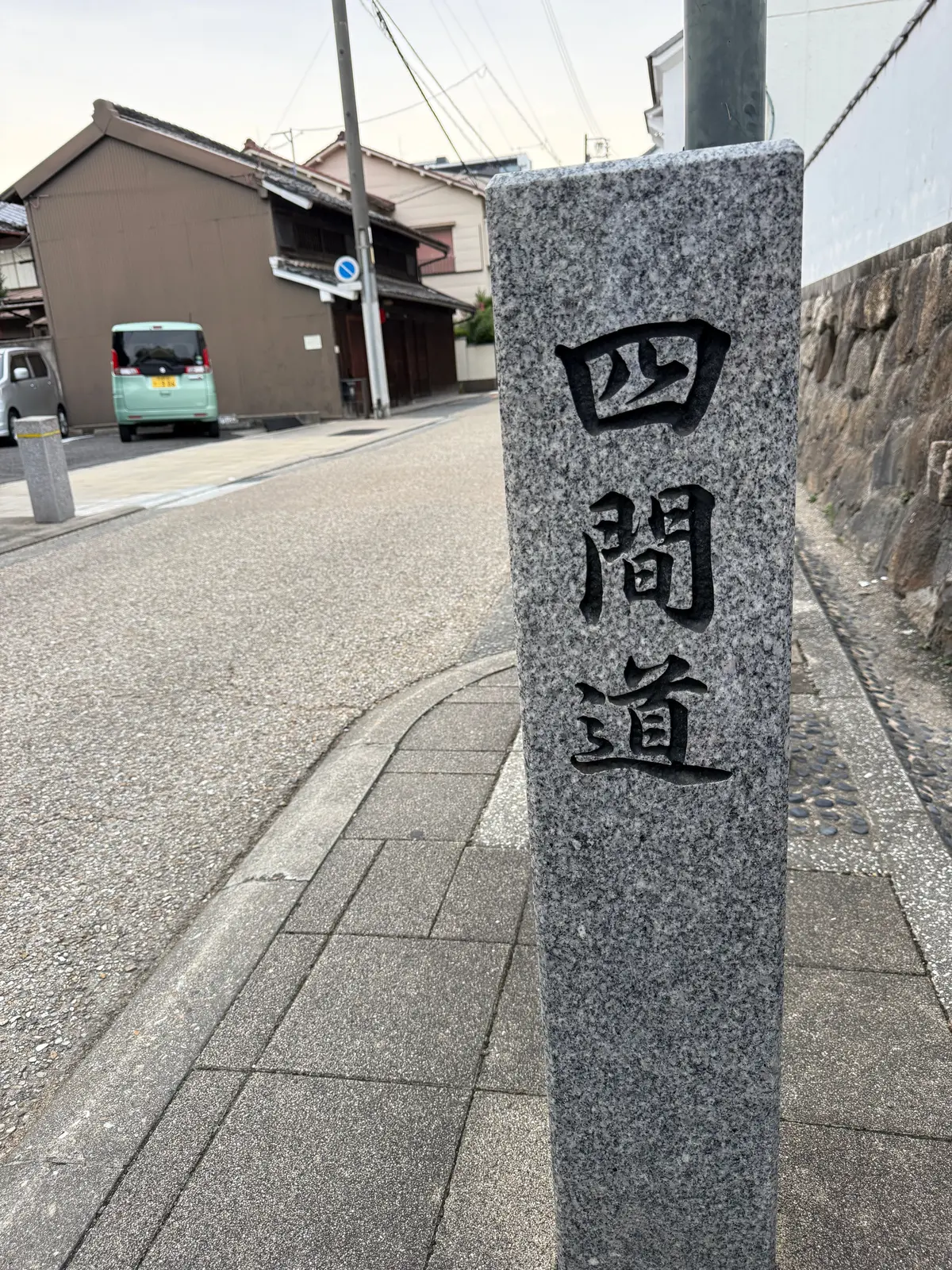 石柱に「四間道」と墨書された標識。背景には、日本の古い民家が並ぶ住宅街の風景。道路、歩道、石垣などが見える。