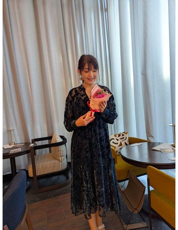 紺色のレースのワンピースを着た女性が、ピンク色の花束を手に笑顔を見せている。背景には白いカーテンとテーブル、椅子が配置された空間。