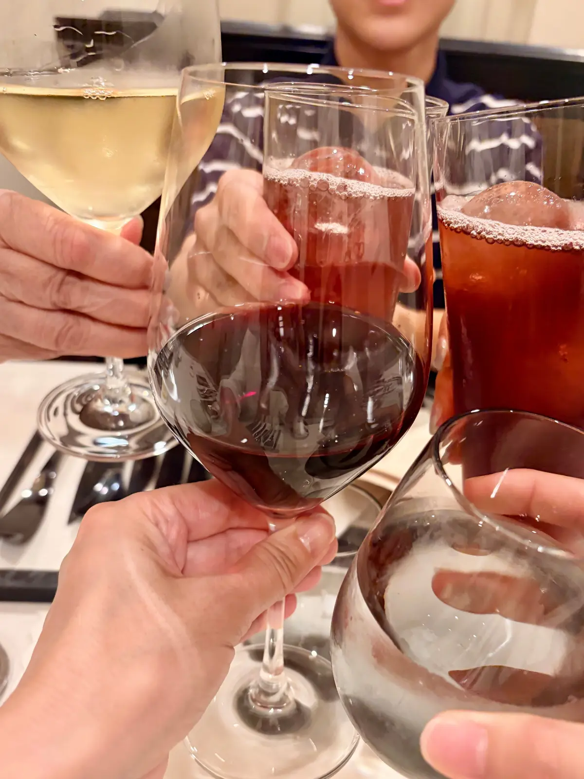 複数のグラスが重なり合って、乾杯の様子。白ワイン、赤ワイン、ロゼワイン、そしてビールが注がれている。背景には人物がぼやけて写っている。