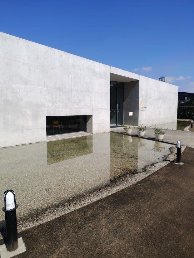 コンクリート造りの長方形の建物。水面が鏡のように建物を反射している。正面には、窓と入り口があり、左右に植栽がある。青空が背景。