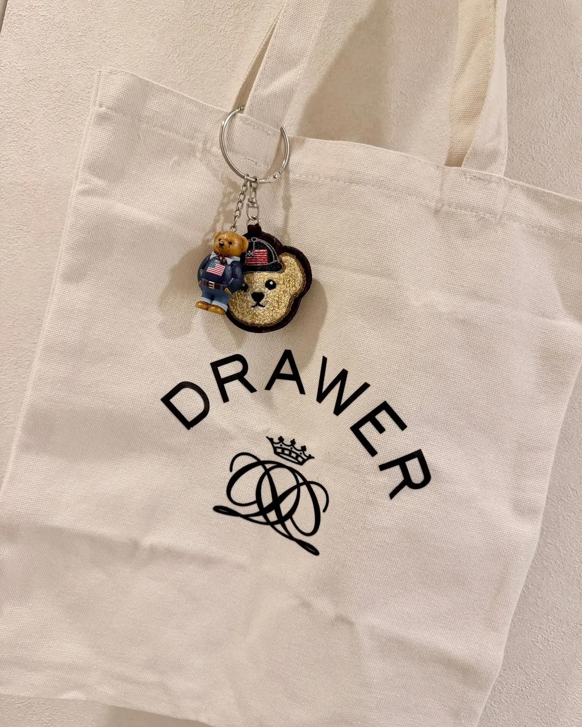 アイボリー色のキャンバストートバッグ。中央に黒色の「DRAWER」の文字と、王冠をかぶった複雑な模様のロゴが配置されている。バッグの持ち手に、アメリカ国旗と帽子をかぶったテディベアのキーホルダーが取り付けられている。