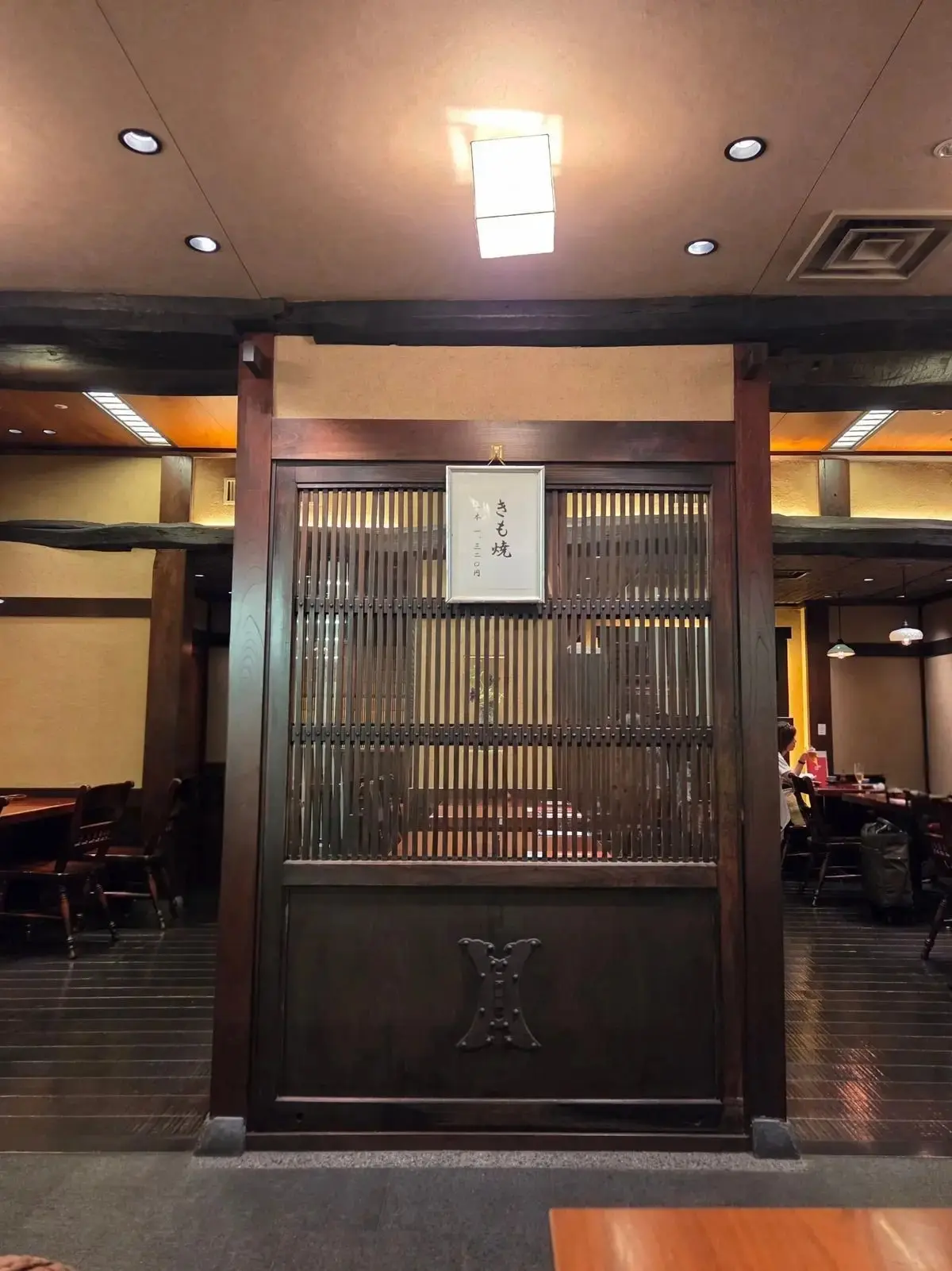 木製の格子戸と、その上に掲げられた白い掛け軸が中央に配置されたレストランの様子。背景にはテーブル席と照明器具が見える。