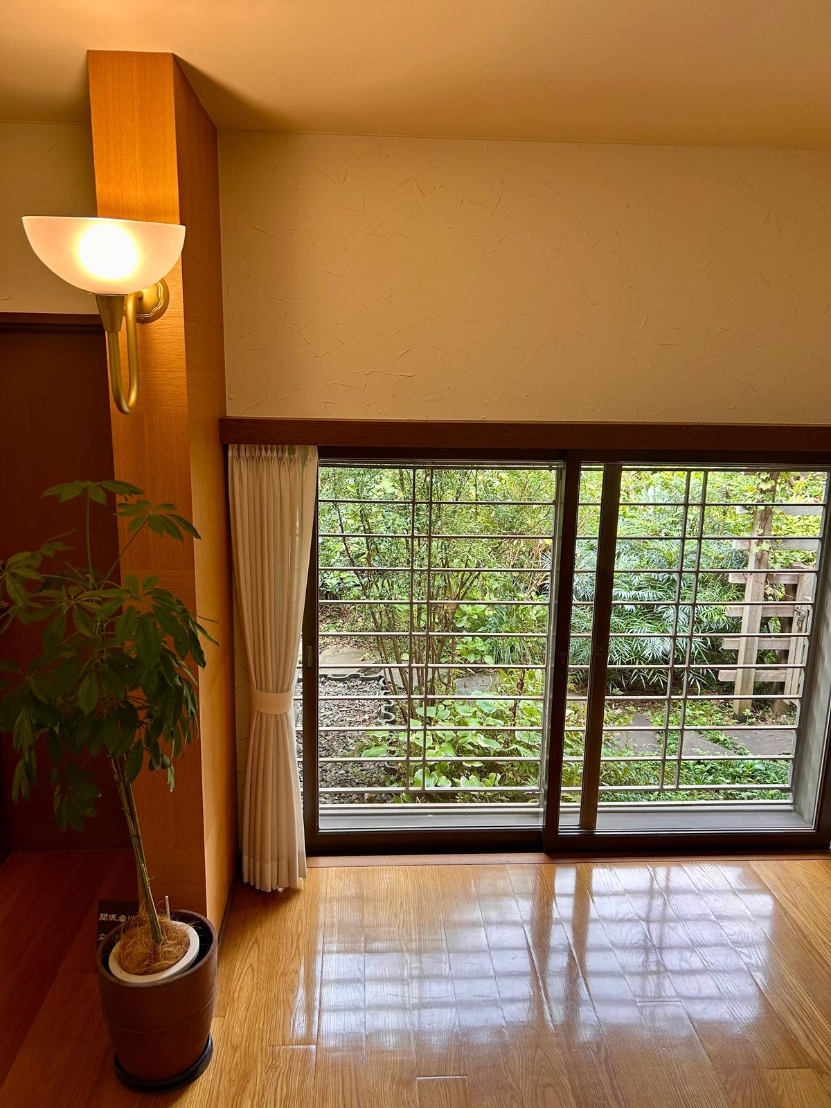 木目調の壁とライト、観葉植物が置かれた部屋。窓の外には緑豊かな庭が見える。窓は格子状で、白いカーテンがかかっている。床は明るい木製。
