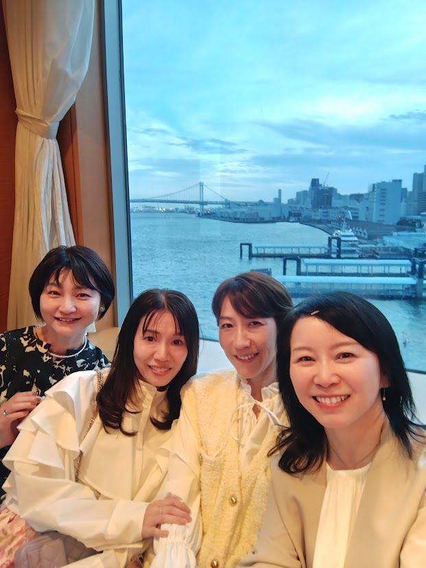 窓から海と橋が見える室内で、4人の女性が笑顔で並んで写っている。女性たちは白っぽいトップスを着用し、左端の女性は黒い模様のトップスを着ている。背景には、青い空と水面、遠くに都市の建物が見える。