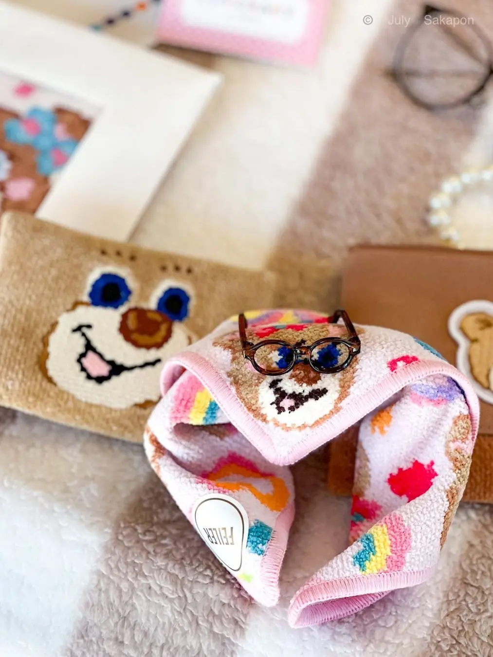 ピンクのタオルにメガネをかけたテディベアのアップ。背景には、チョコレートの箱や化粧品、その他ギフトらしき品々が置かれている。