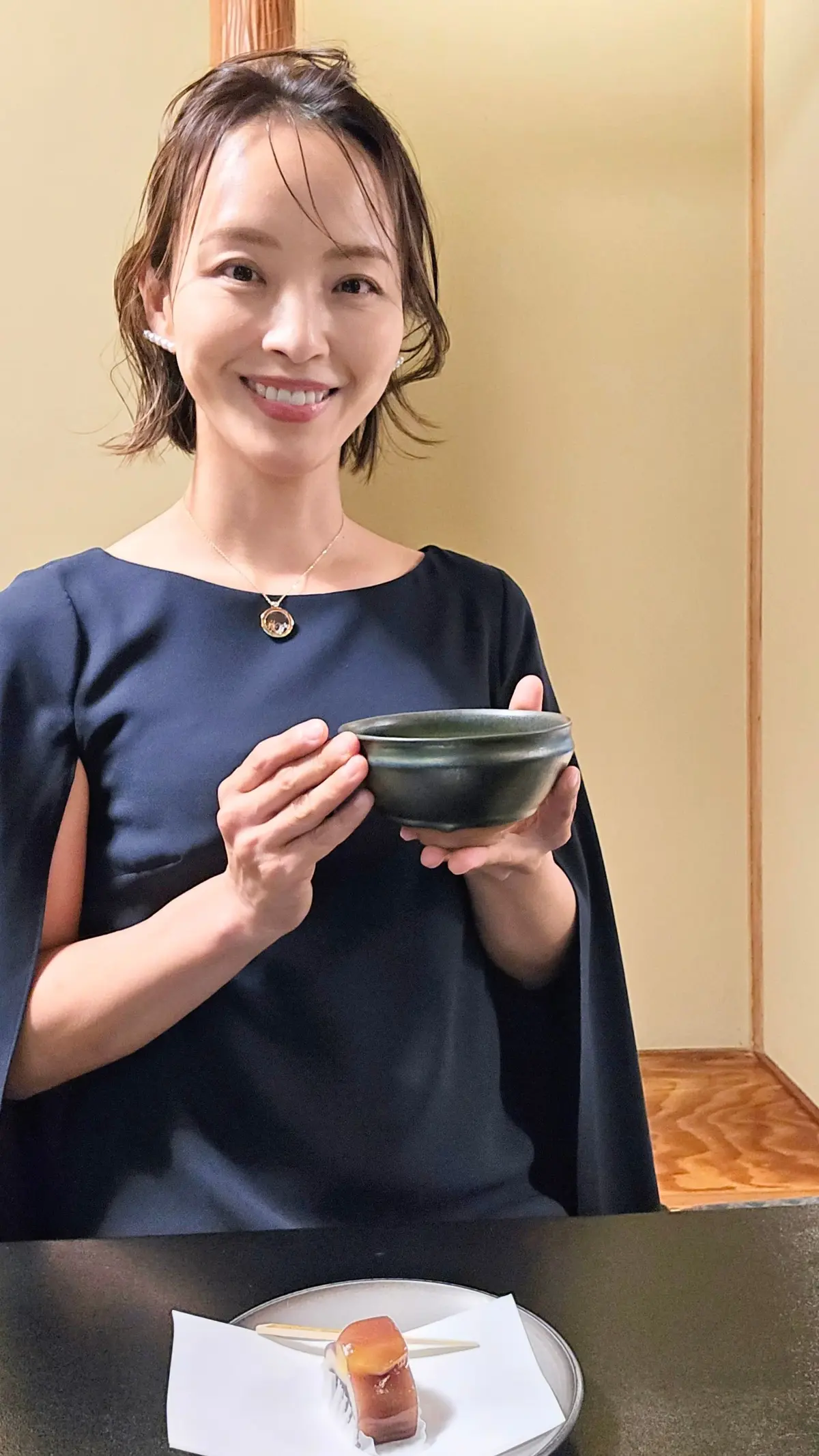 笑顔の女性が、緑色の陶器の碗を持ち、テーブルの上の寿司の横に立っている。紺色のワンピースを着ており、首には小さなペンダントを付けている。背景は淡い黄色。
