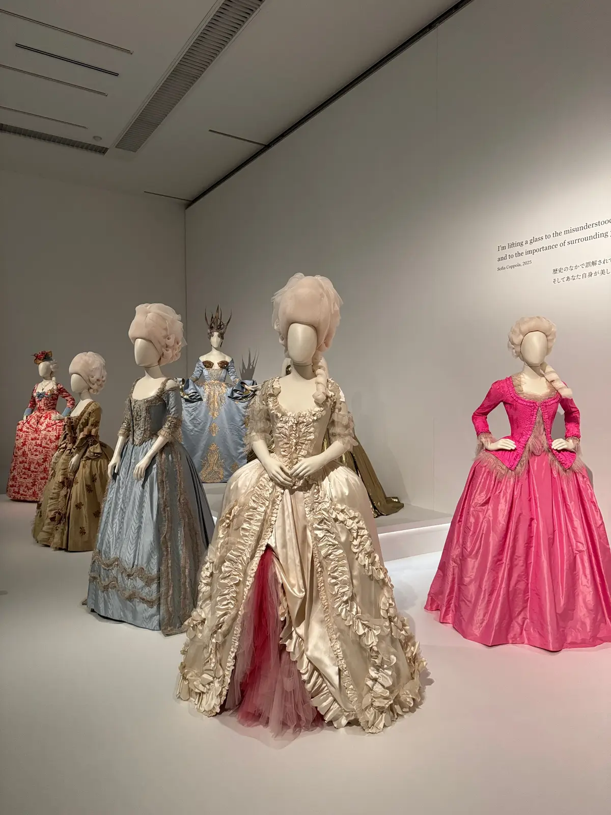 展示ケースに並んだ、18世紀風の豪華なドレス5着。クリーム色、水色、ピンク、赤、緑のドレスが、白髪の付け毛をつけたマネキンに着せられている。フリルやリボン、レースなどの装飾が施され、豪華な雰囲気を醸し出している。
