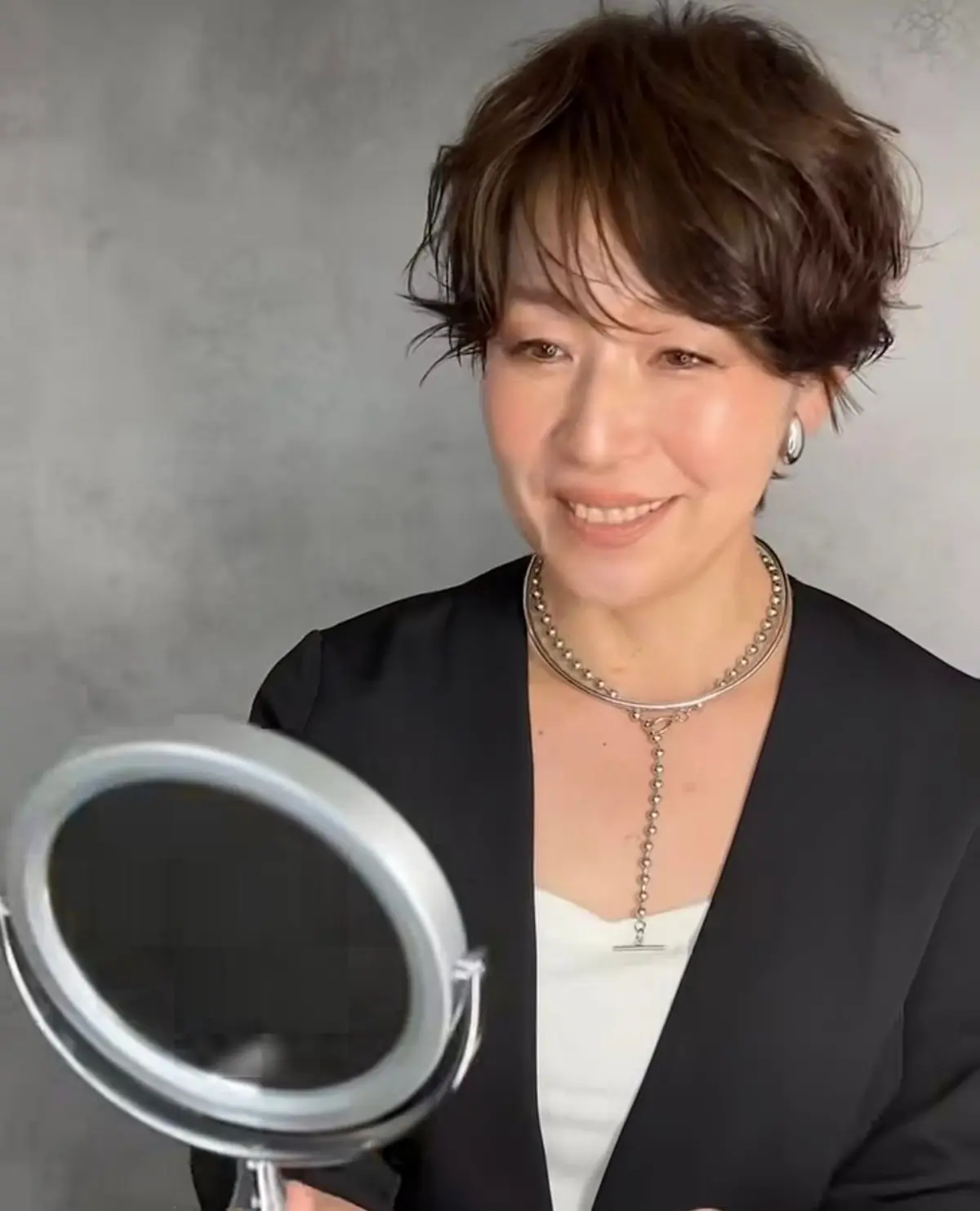 笑顔の女性が鏡を見ている。茶色の短髪で、黒のジャケットと白いインナーを着用。シルバーのネックレスを2連で身につけている。背景はグレーの無地。