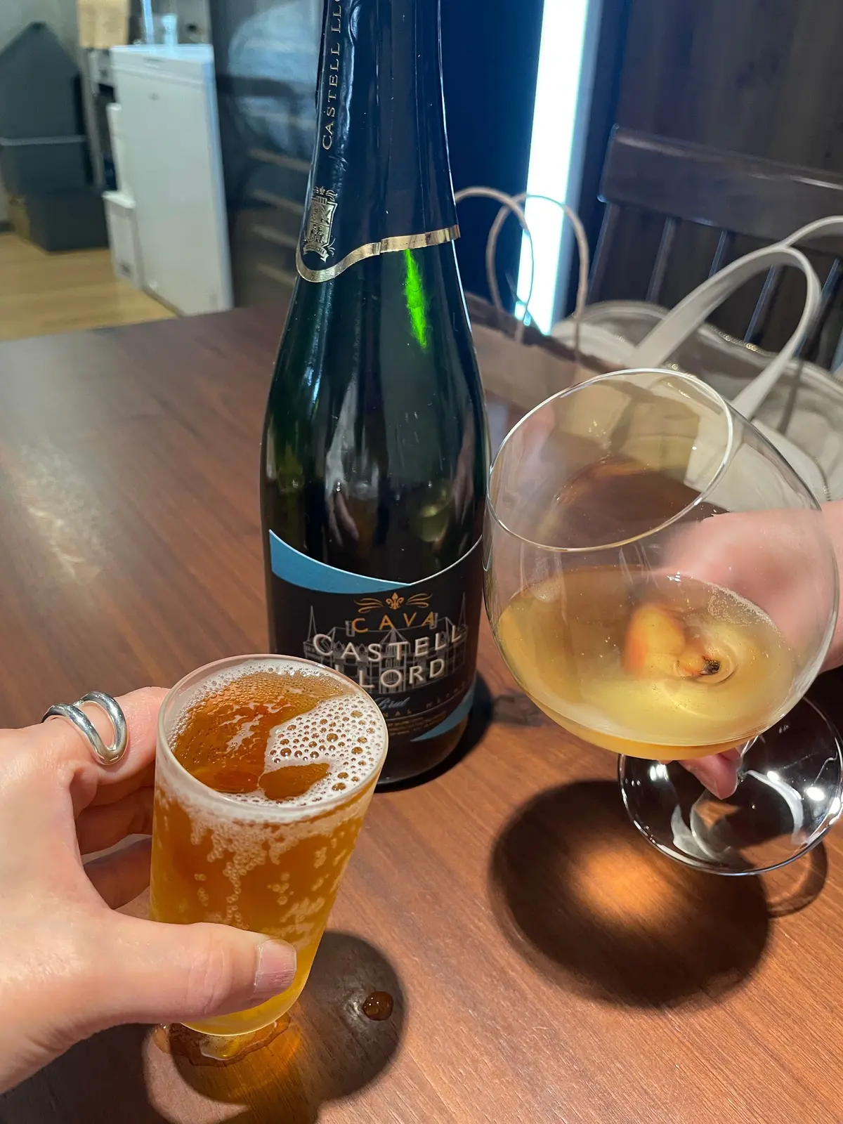 木製テーブルにシャンパンのボトルと二つのグラスが置かれている。左のグラスは泡立つビールで、右のグラスはオレンジ色の飲み物と果物が入っている。