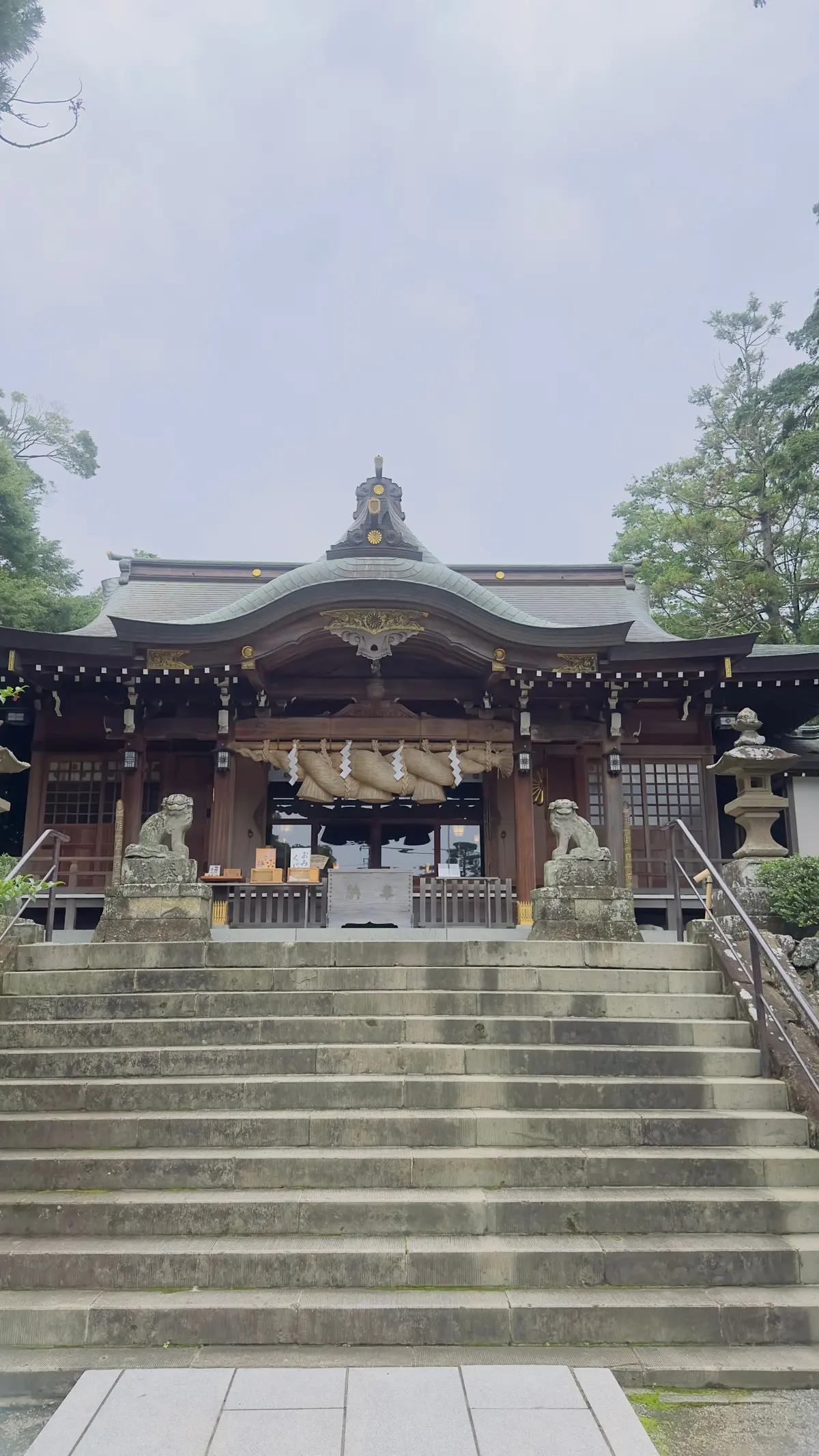 石段を上った先にある伝統的な神社。木造の建物に石の狛犬が配置され、正面には提灯が吊るされている。緑豊かな木々が背景に広がる。