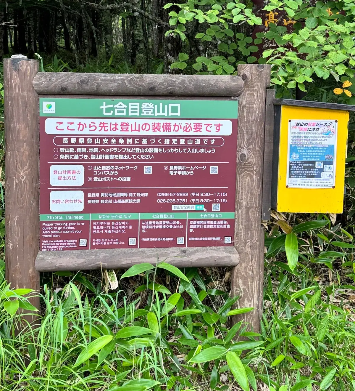 緑豊かな木々に囲まれた山道脇に、木製の標識が設置されている。標識には日本語で「七合目登山口」と大きく書かれ、登山に関する注意事項や登山届の提出方法などが記載されている。右側には山の注意に関する掲示板が設置され、背景にはさらに緑の木々が広がっている。