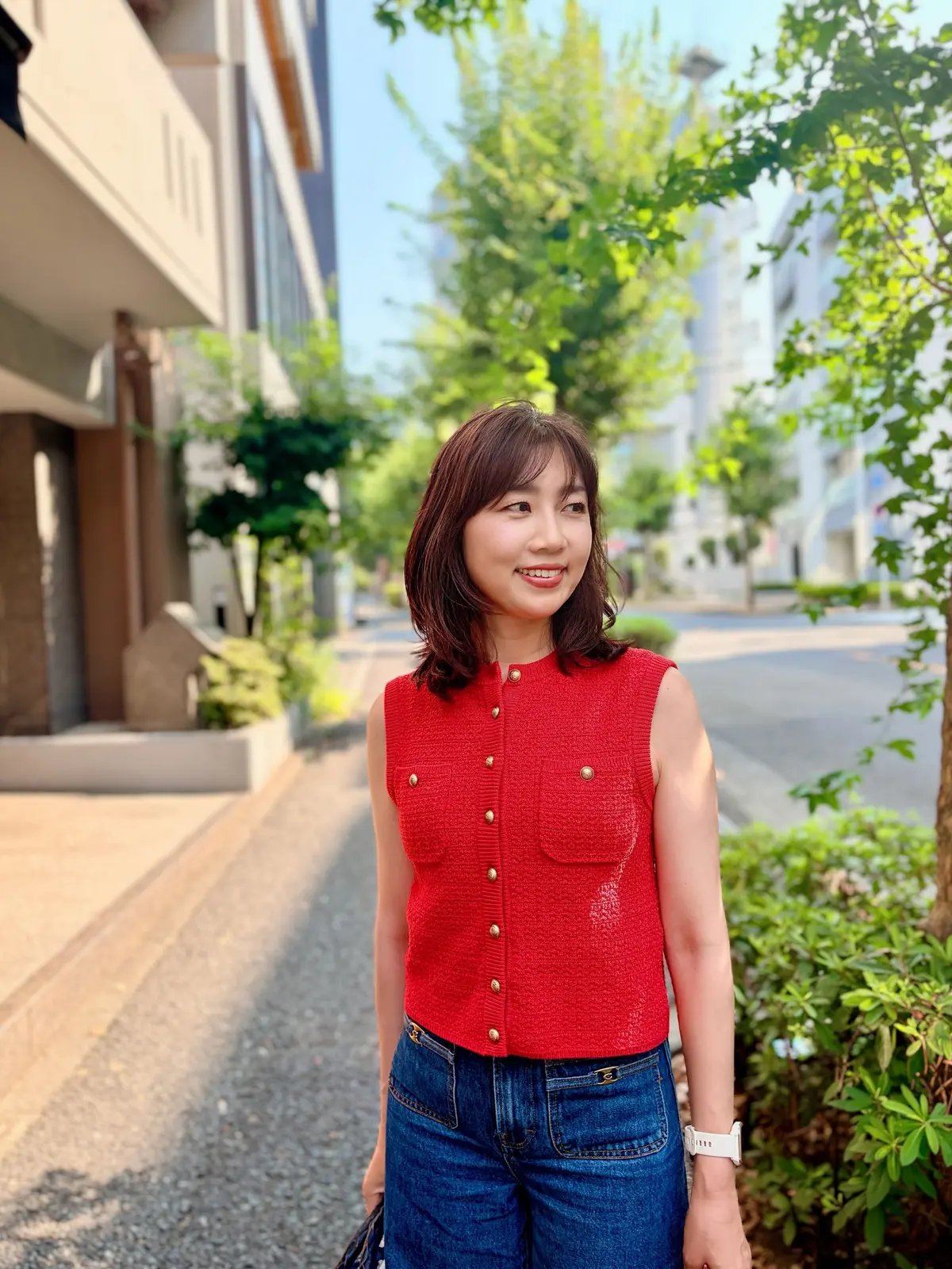 赤いニットのトップスにデニムを着用した女性が、街並みを背景に笑顔を見せている。右腕に白いスマートウォッチを着用。
