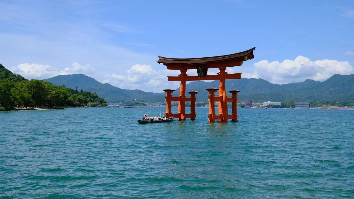 瀬戸内海の穏やかな水面の中央に、鮮やかなオレンジ色の鳥居が浮かぶ。背景には緑豊かな山々が連なり、青空に白い雲が浮かんでいる。手前には小さな船が航行している。