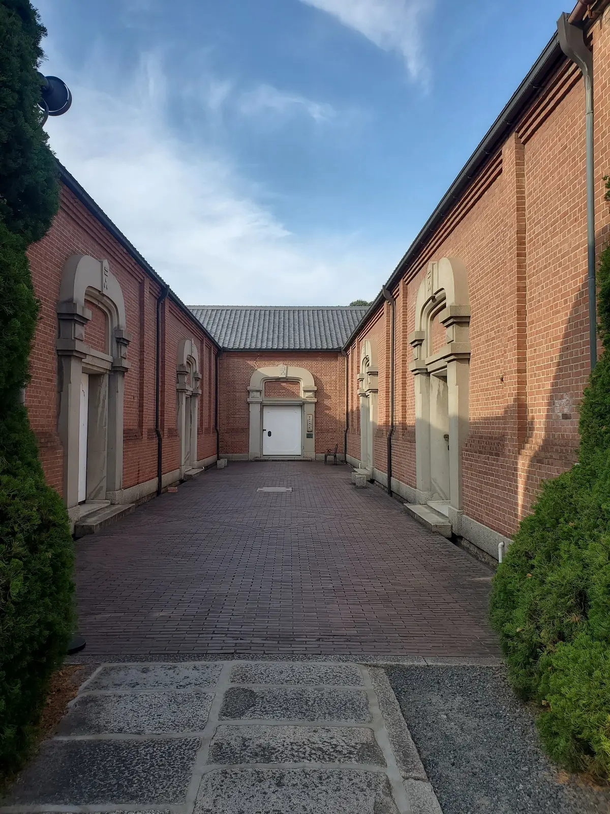 赤レンガの建物が左右に並び、中央に通路が続く風景。通路は赤茶色のタイルで舗装され、遠くに白いドアが見える。青空に白い雲が浮かんでいる。