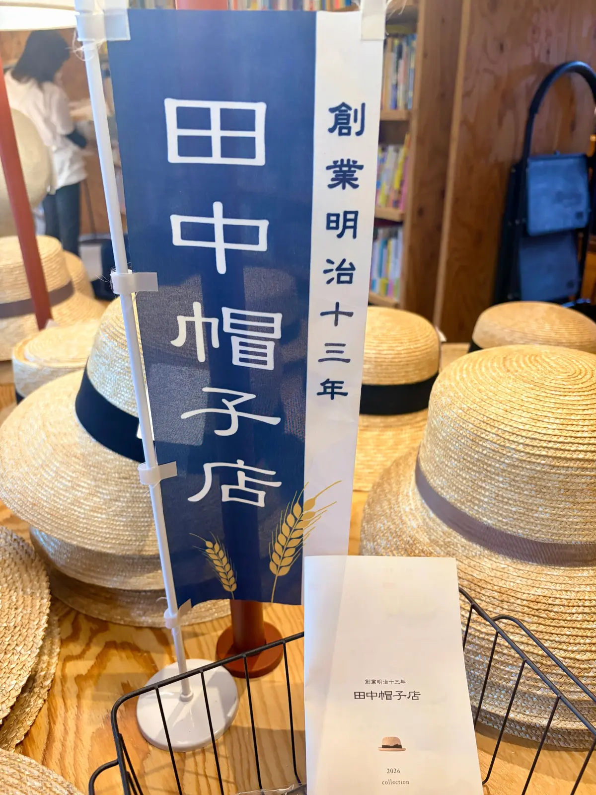 縦長の看板と麦わら帽子が置かれた店内。看板は青と黄色の配色で、中央に「田中帽子店」の文字が大きく書かれている。下部には麦の穂のイラスト。手前に黒いワイヤーバスケットに入った麦わら帽子が並んでいる。