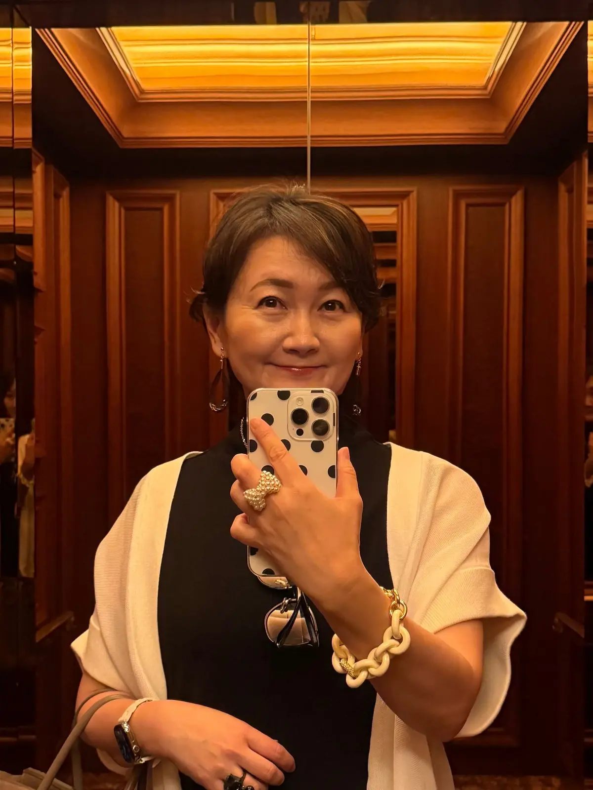 黒いネックホルダーに白いシャツを羽織った女性が、スマートフォンで自撮りしている。背景は木目調の豪華な内装。女性は笑顔で、指には大きな指輪、腕にはブレスレットを身につけている。