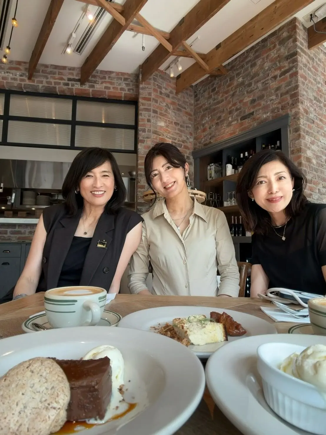 カフェでデザートを楽しむ3人の女性。左から黒の服、中央はベージュのシャツ、右は黒のワンピース。それぞれコーヒーやケーキ、生クリームを前に笑顔。背景にはレンガの壁と大きな窓がある。