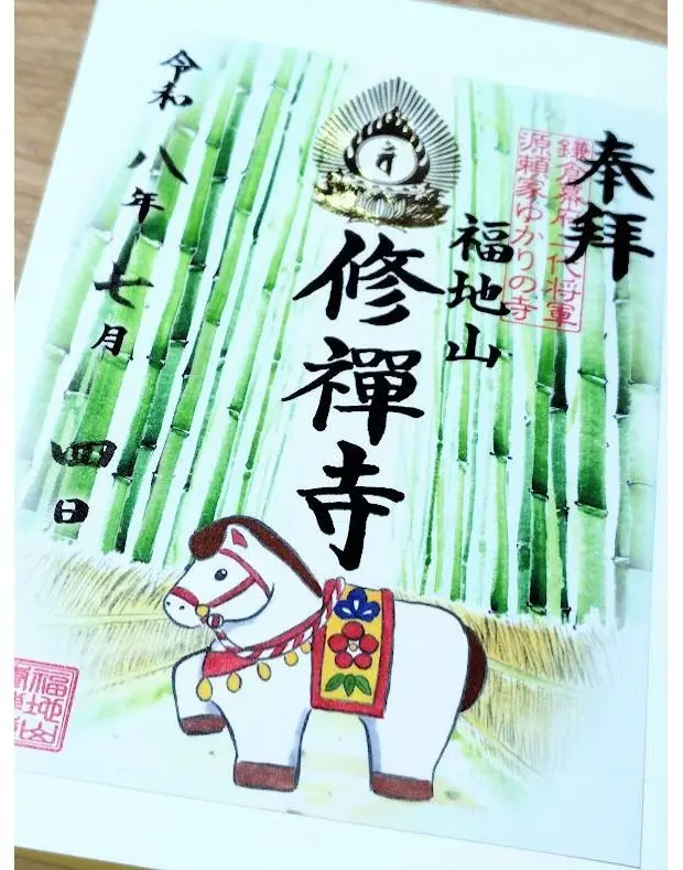 緑竹の背景に、白地に黒と赤の文字で「修禅旅行寺」と書かれた書道作品。中央上には仏像、下部には白馬の絵馬が描かれている。右下に朱色の印章。