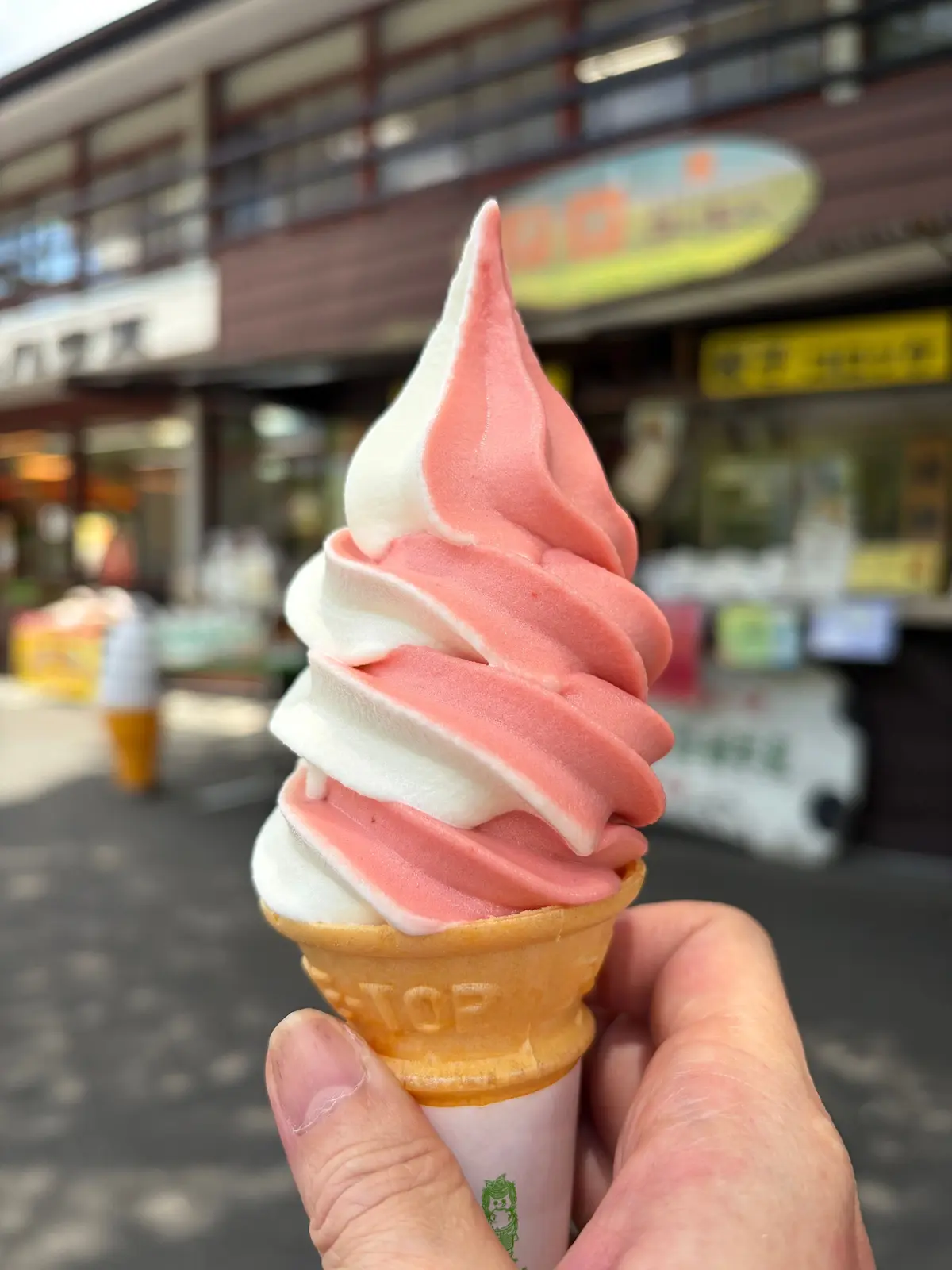 ピンクと白のアイスクリームがコーンに盛られた写真。手で持たれ、背景には店構えが見える。