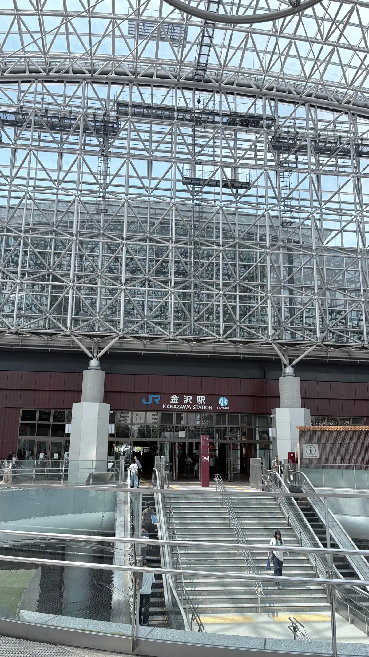 金沢駅の駅舎。複雑な鉄骨構造の屋根が特徴的で、正面にはJRのロゴと駅名標がある。エスカレーターと階段で人が移動している。