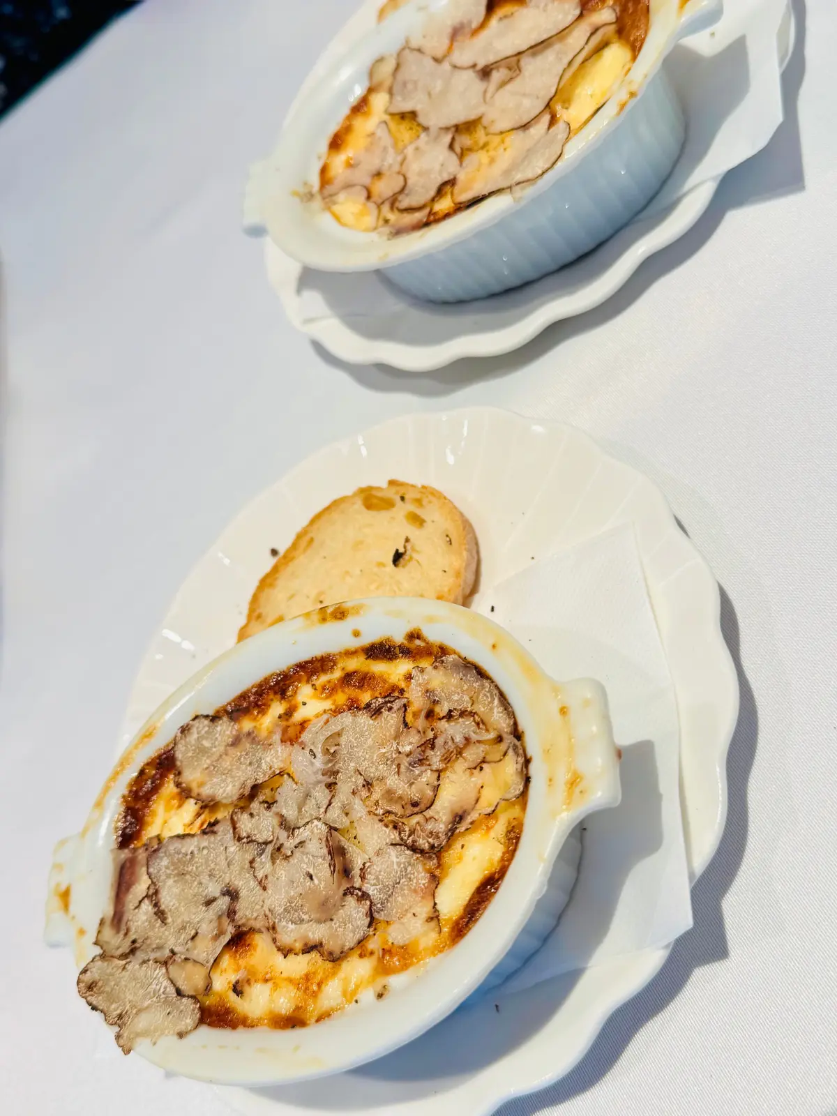 白い皿に盛り付けられた、焦げ目がついたクリーム色の器に入った料理。器の上には薄切りの肉が乗っており、横には丸いクッキーが添えられている。