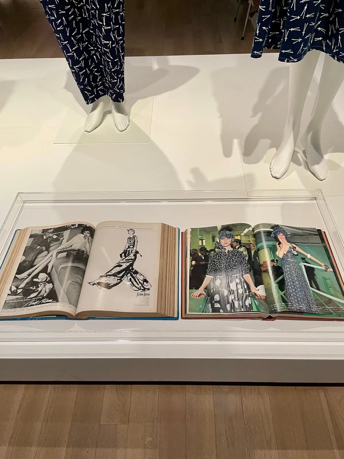 白い台に置かれた大型のファッション雑誌。左ページには白黒写真、右ページにはカラー写真が掲載。背景には、青と白の柄のパンツを履いたマネキンが配置されている。