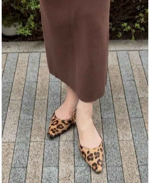 茶色のミモレ丈スカートを履いた女性の足元。茶色のタイツと、それぞれ異なる柄のレオパード柄のフラットシューズを履いている。背景には石畳の道と、緑の植物が見える。