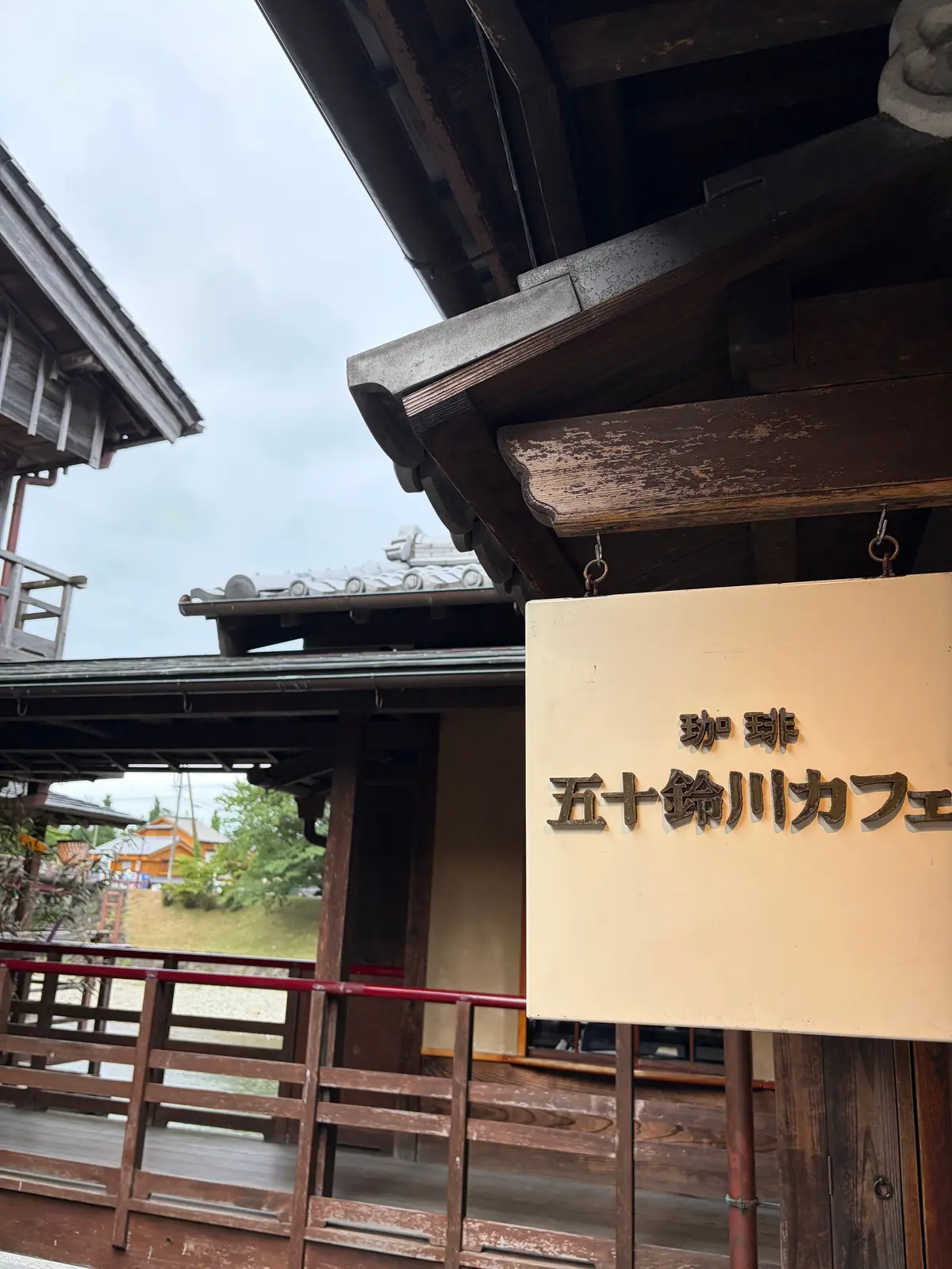 木造建築の軒下にある、ベージュ色の看板。「弥陀寝 五十鈴川カフェ」と黒文字で表記されている。手すり付きの木製テラスが前景に見える。背景には緑の木々が茂り、曇り空が広がっている。