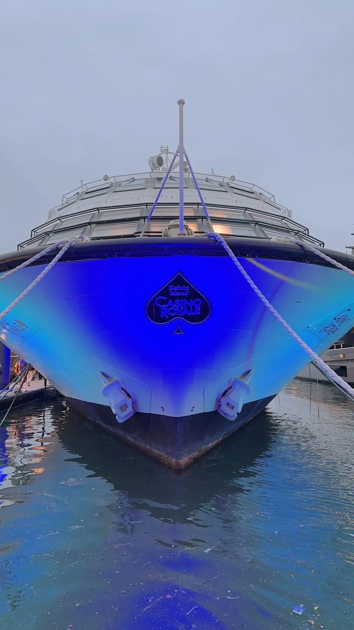 青いライトに照らされたクルーズ船の船首。中央には「Casino Royale」のロゴが配置され、水中に係留索が垂れている。背景は薄曇りの空。