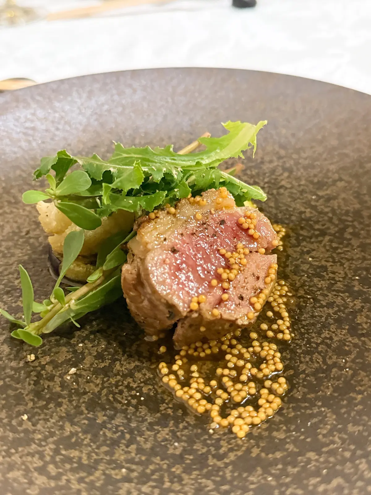 茶色の皿に盛り付けられた、中心にピンク色の肉、周りに黄色い粒状ソース、緑の葉野菜が添えられた料理。