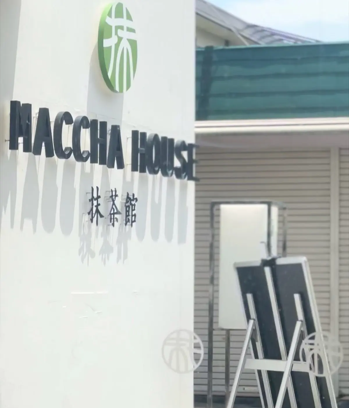白い壁に「HACCHA HOUSE」と大文字で書かれた看板、緑のロゴと日本語の表記があり、屋外に置かれた黒いボードが並んでいる