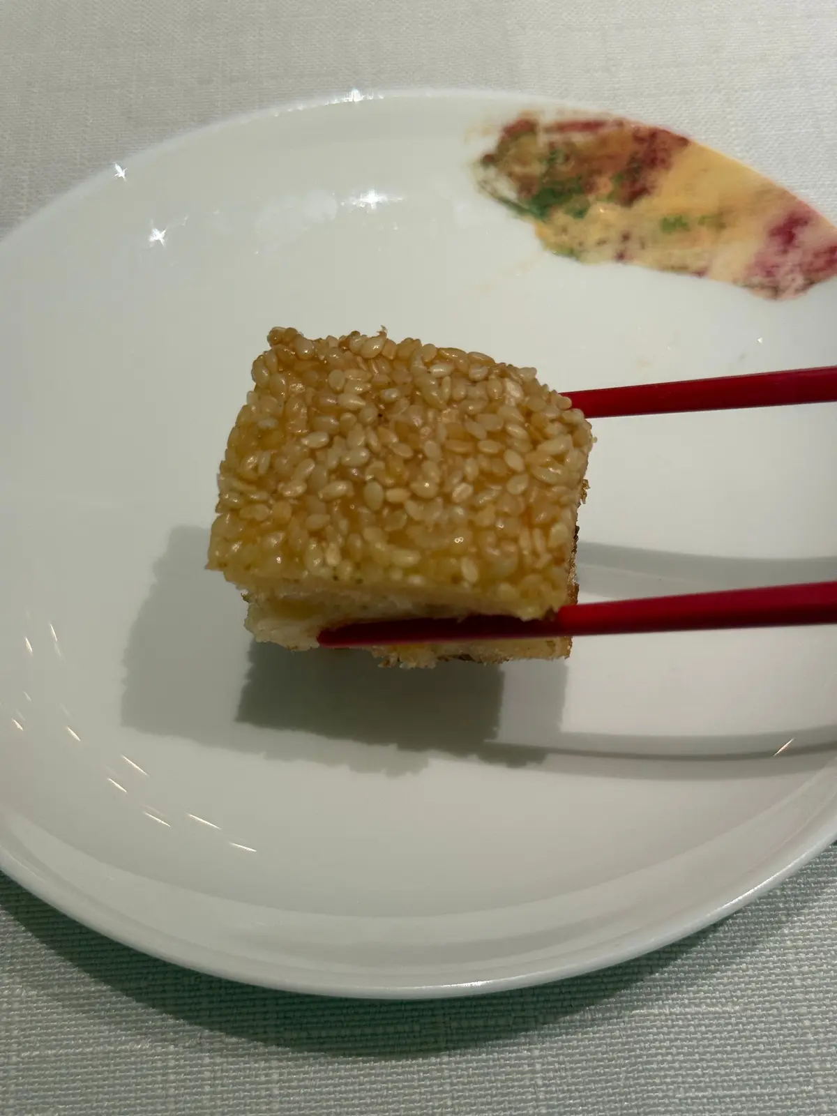 白い皿に赤い箸で挟まれた、白ごまをまぶした四角いお菓子。背景には淡いピンク色のソースが塗られたものが少し見える。