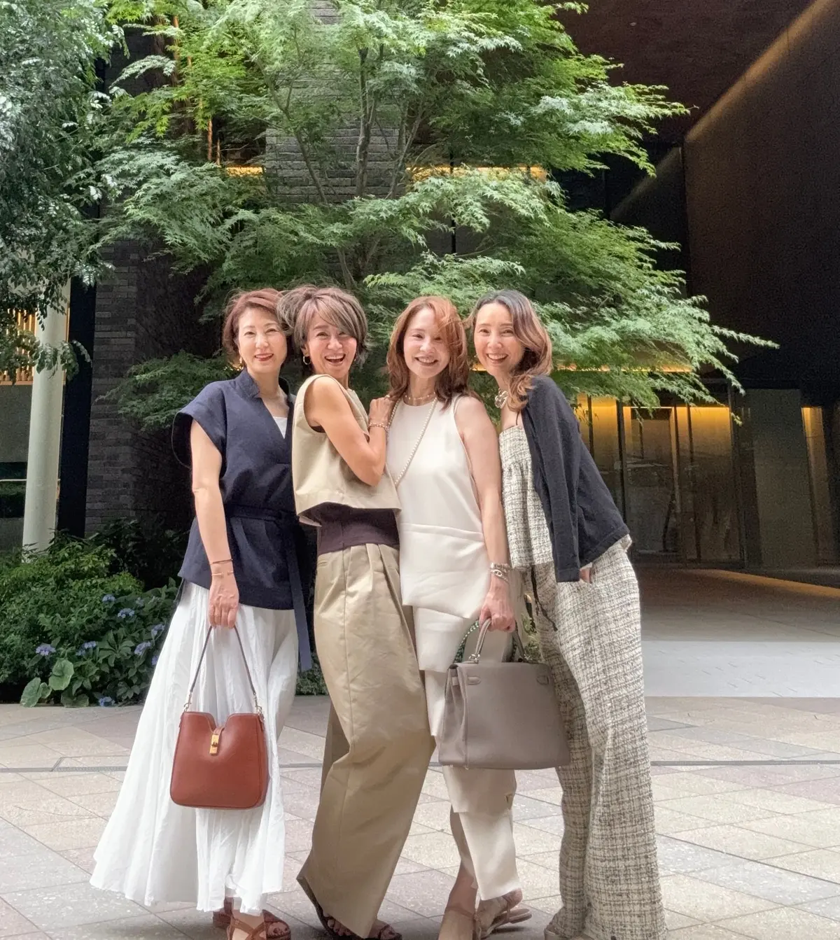 4人の女性が並んで明るく笑い、屋外の建物前でポーズをとっている。手荷物を持った女性が、緑豊かな木々が背景に広がる。