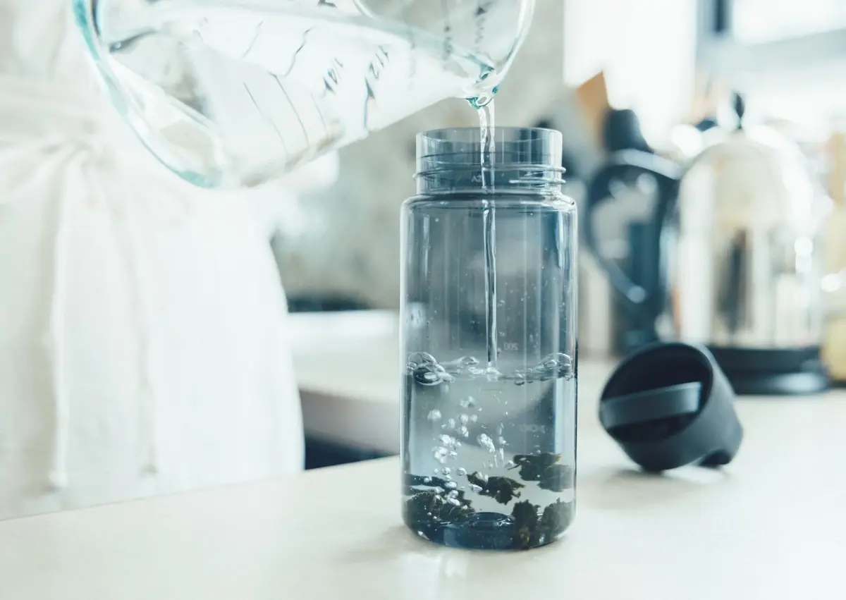 透明な水差しから水が注がれる様子。水差しの中には水が満たされ、注ぎ口から細い水流が透明なボトルに落ちている。ボトルの底には茶色い粉末状のものが沈殿しており、お茶を淹れている状況がわかる。
