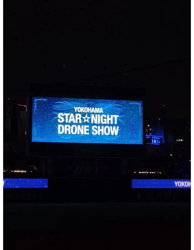 夜の暗闇に浮かぶ大型ビジョン。横浜の夜空を彩る「STAR NIGHT DRONE SHOW」の文字が青色で輝いている。下部にはコカ・コーラや「DRY」のロゴ、その他の広告が表示されている。