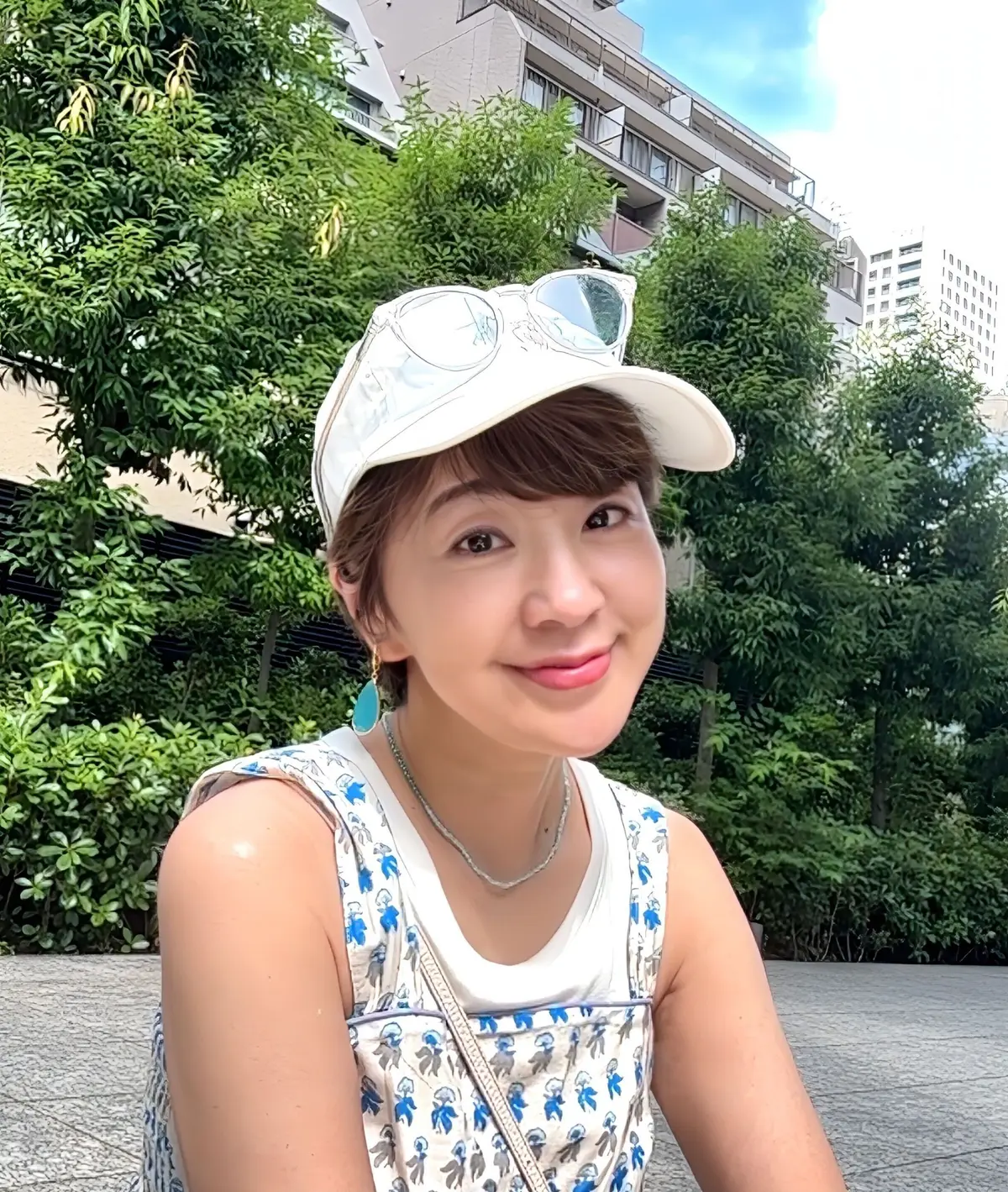 白いキャップを被った女性が、緑豊かな木々と高層ビルを背景に笑顔を見せているセルフィー。青色の柄のトップスを着ており、ネックレスとイヤリングを身につけている。