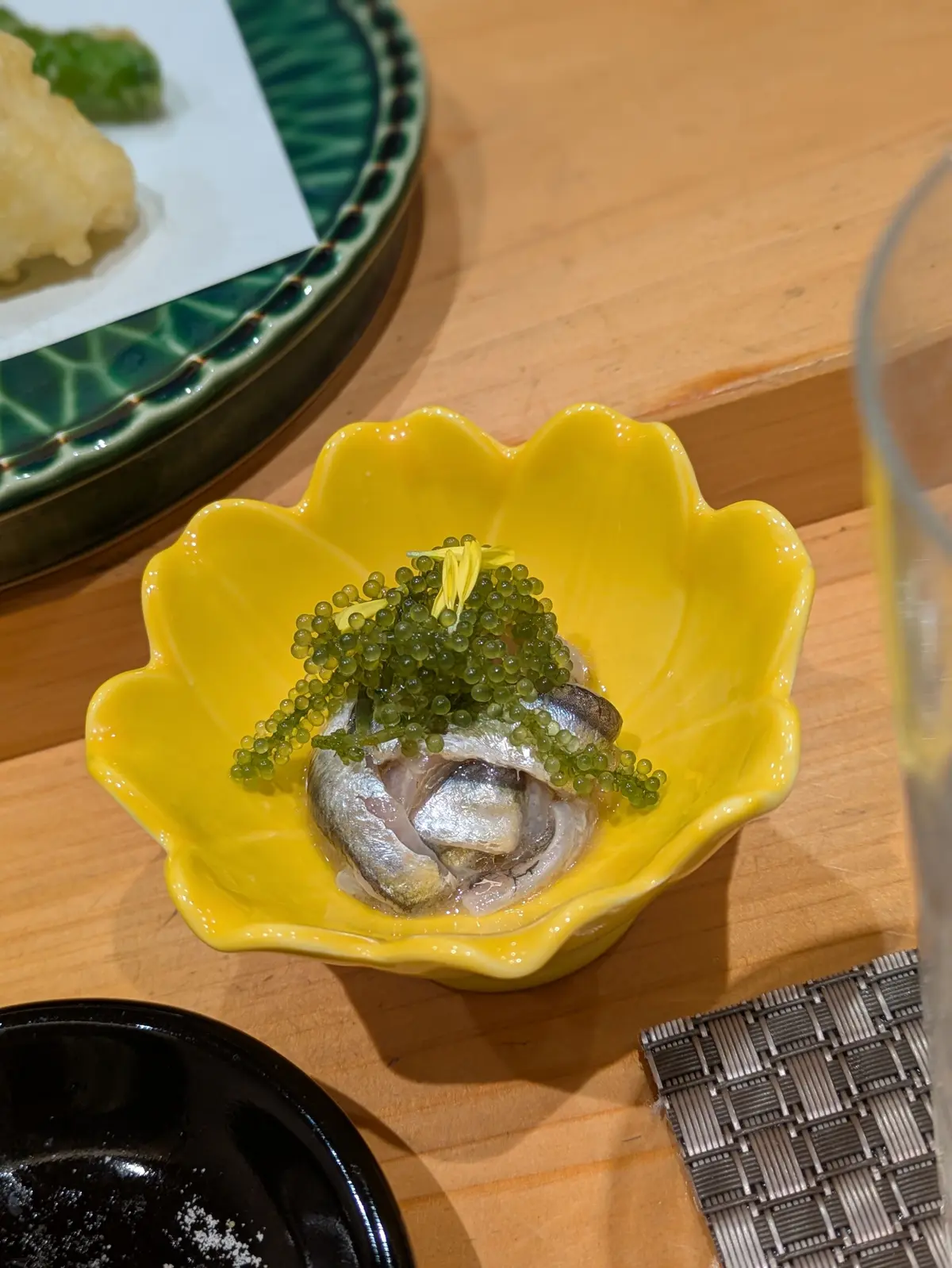 黄色の花びら型の器に、銀色のカツオの腹身と緑のワサビのつぶが乗せられ、小さな黄色い花が添えられている