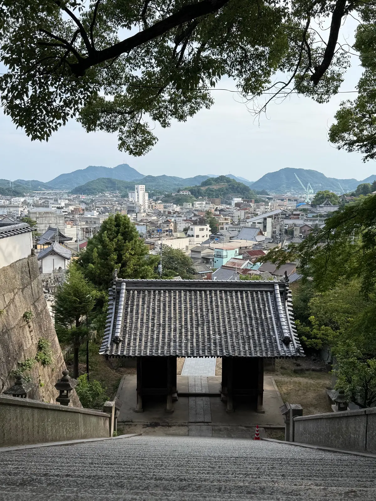 石畳の階段を上がると、伝統的な屋根の門が現れ、その先には都市の風景と遠くの山々が広がる。緑の葉が画面上部を覆っている。