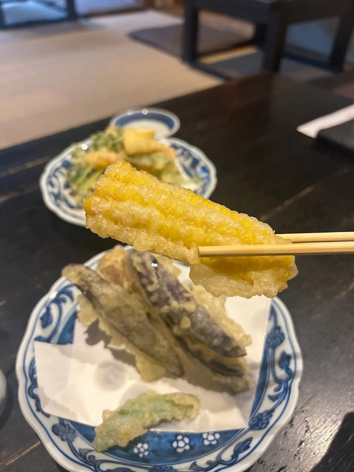 天ぷら手前にはコーンの天ぷらが箸で支えられ、奥には野菜の天ぷらが並んでいる。